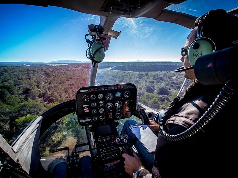 Pilote d'hélicoptère en vol, vu de l'intérieur du cockpit avec tableau de bord et paysage forestier en arrière-plan.