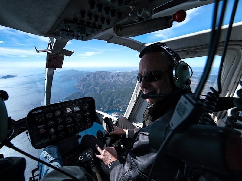 Pilote souriant portant casque et lunettes de soleil, aux commandes d'un hélicoptère en vol au-dessus d'un paysage montagneux.
