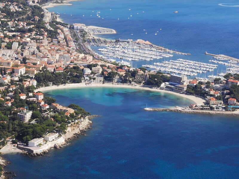 Vue aérienne d'une baie avec plage, port de plaisance et habitations en bord de mer sous un ciel clair.