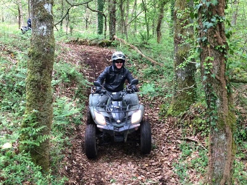 Homme souriant conduisant un quad sur un chemin forestier couvert de feuilles et entouré d'arbres.