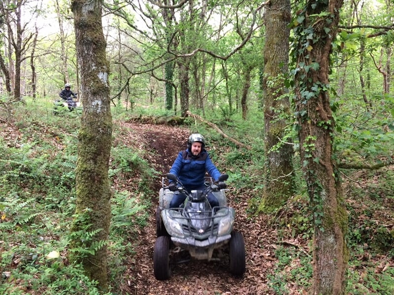 Personne en blouson bleu conduisant un quad sur un sentier boisé avec des arbres verts autour.