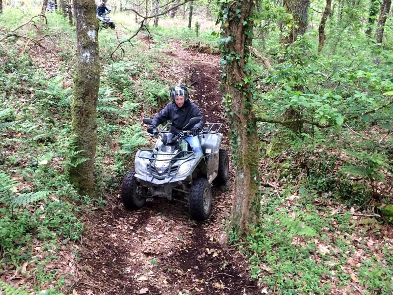Homme en tenue noire conduisant un quad sur un chemin forestier avec des feuilles mortes au sol.