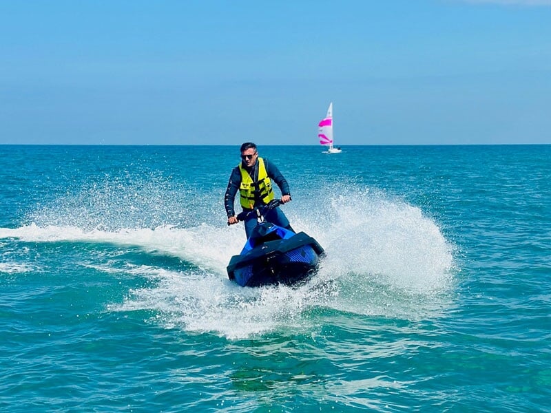 Un homme dirigeant son jet-ski bleu en ligne droite sur l'eau, créant une grande vague derrière lui. Il porte un gilet de sauvetage jaune et se dirige vers l'avant avec assurance. En arrière-plan, un