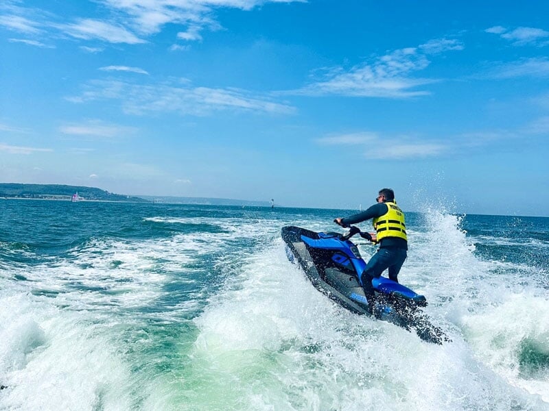Homme effectuant un saut spectaculaire avec son jet-ski bleu, laissant derrière lui une traînée d'écume blanche. Le ciel est dégagé et bleu, et le paysage côtier à l'horizon est visible, accentuant la