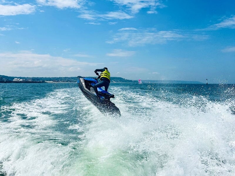 Le jet-ski bleu est capturé en plein saut au-dessus des vagues, avec l'homme en gilet de sauvetage jaune accroché fermement au guidon. La mer s'étend à perte de vue sous un ciel bleu clair, offrant un