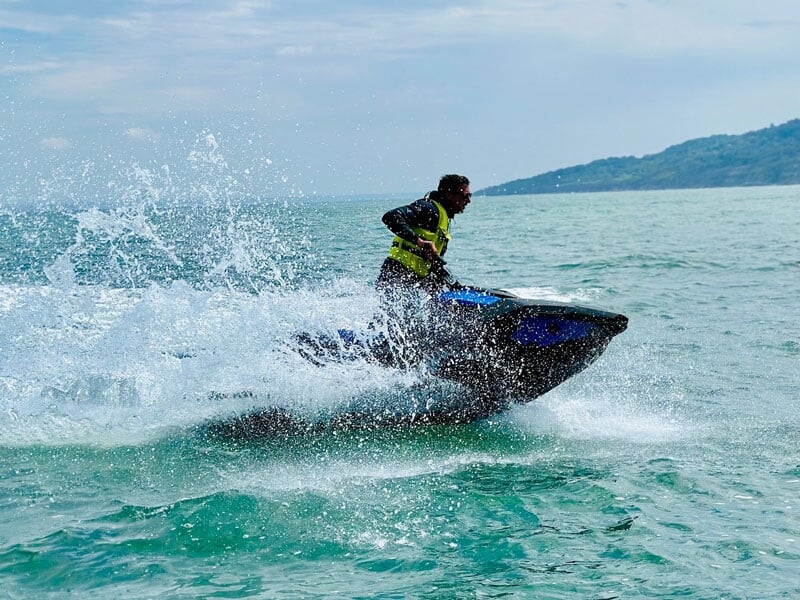 Un jet-ski bleu traverse la mer, projetant de grandes gerbes d'eau alors qu'il effectue un virage serré. Le pilote, vêtu d'un gilet de sauvetage jaune, semble totalement maîtriser l'engin, tandis que