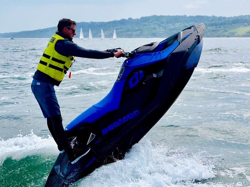 Homme en équilibre sur un jet-ski bleu, en train de réaliser une figure avec l'avant du véhicule levé hors de l'eau. Il porte un gilet de sauvetage jaune et semble concentré. À l'arrière-plan, on dist