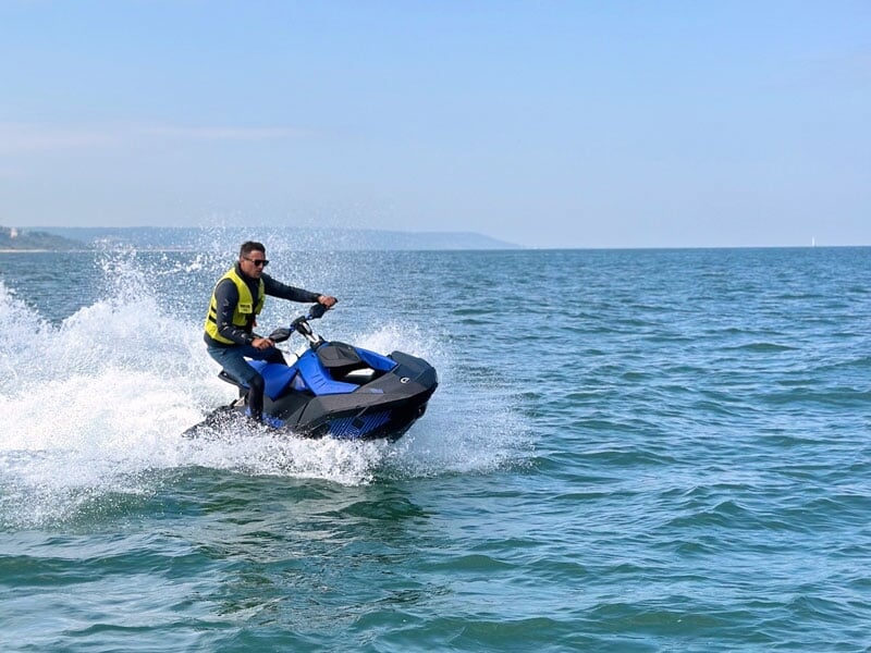 Homme conduisant un jet-ski bleu à grande vitesse sur une mer calme. Des éclaboussures d'eau l'entourent alors qu'il file vers l'avant, vêtu d'un gilet de sauvetage jaune et de lunettes de soleil. Le