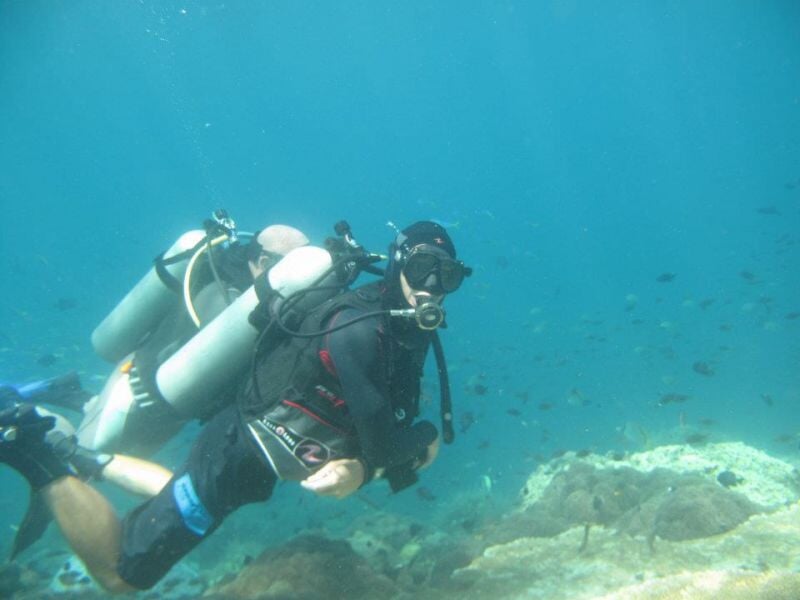 Plongeur équipé de bouteilles d'air explore un récif sous-marin avec des poissons dans une eau claire et bleue.