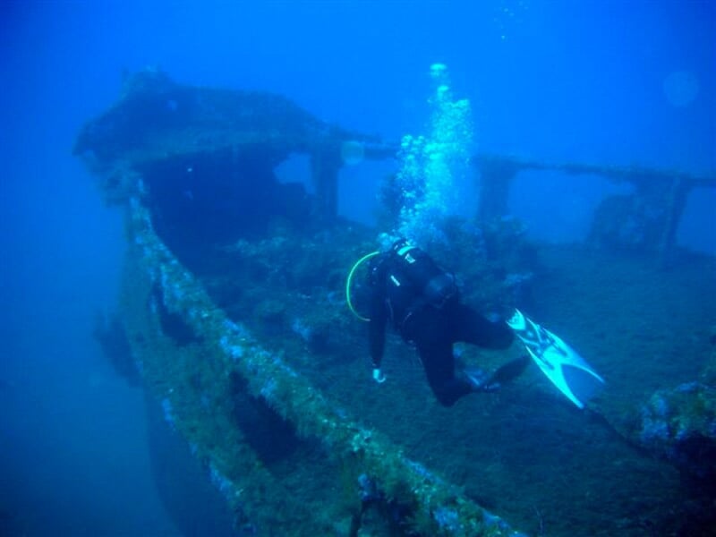 Plongeur en combinaison noire explorant une épave sous-marine recouverte d'algues.