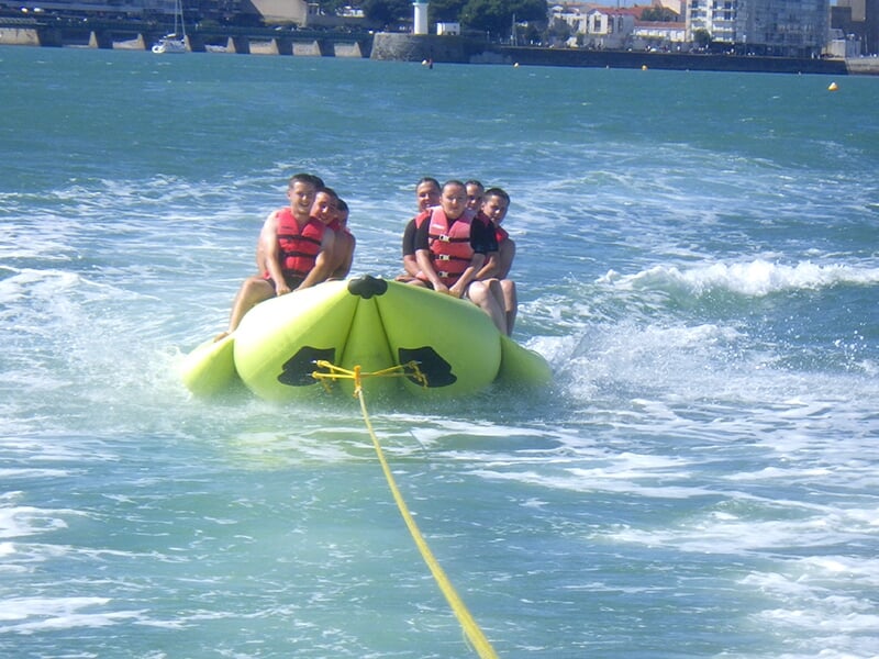 Un groupe de six personnes portant des gilets de sauvetage rouges est assis sur une bouée gonflable banane verte, tractée sur l’eau, avec un décor urbain en arrière-plan.