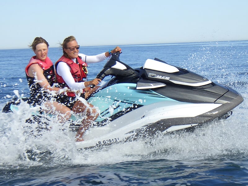 Deux femmes portant des gilets de sauvetage rouges profitent d’une balade en jet ski blanc et bleu, traversant les vagues avec des éclaboussures d’eau sous un ciel dégagé.