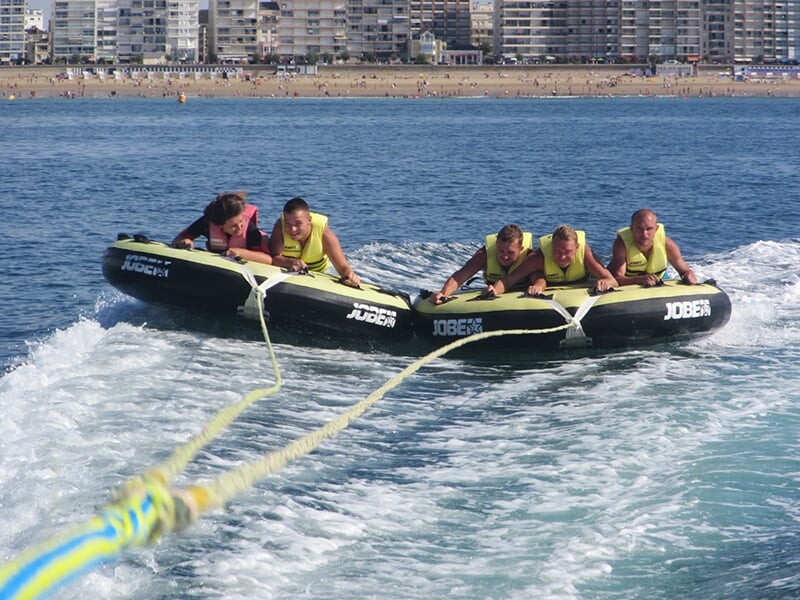Quatre jeunes hommes en gilets de sauvetage jaunes sont allongés sur une grande bouée gonflable noire de marque Jobe, agrippés fermement alors qu’ils sont tirés à pleine vitesse sur la mer, avec une p