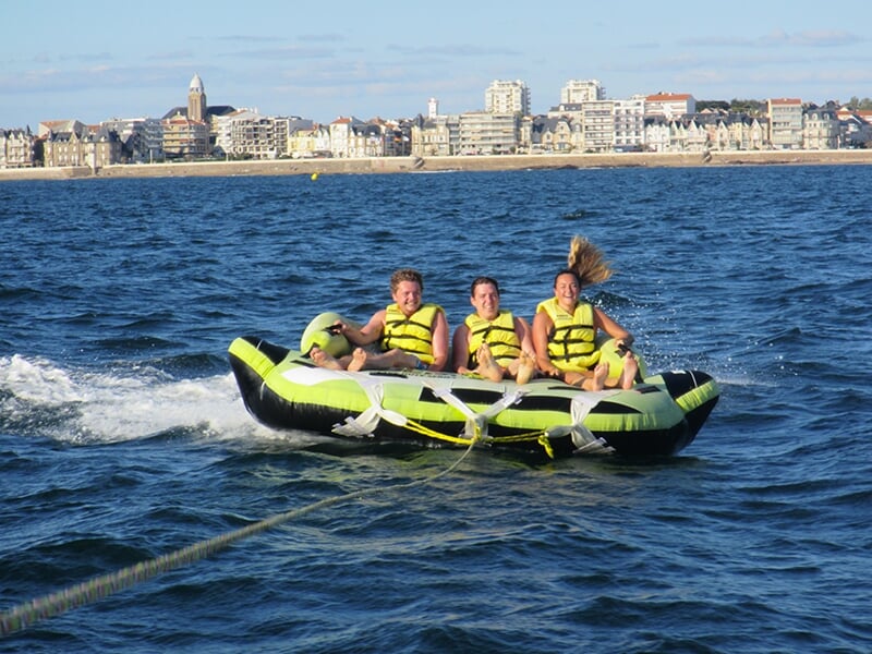 Trois personnes portant des gilets de sauvetage jaunes rient et profitent de leur balade sur une bouée gonflable verte et noire, tirée sur une mer légèrement agitée, avec une ville côtière en arrière-