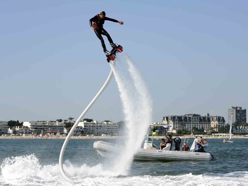 Personne en combinaison noire réalisant une figure acrobatique en flyboard au-dessus de l'eau près d'une plage urbaine.
