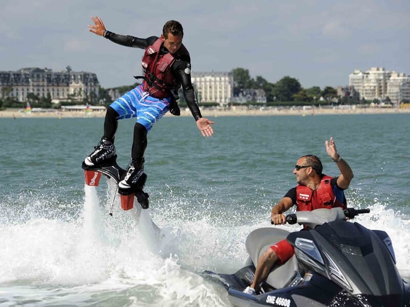 Homme en short bleu sur un flyboard saluant un autre homme sur un jet ski dans une mer calme près du rivage.