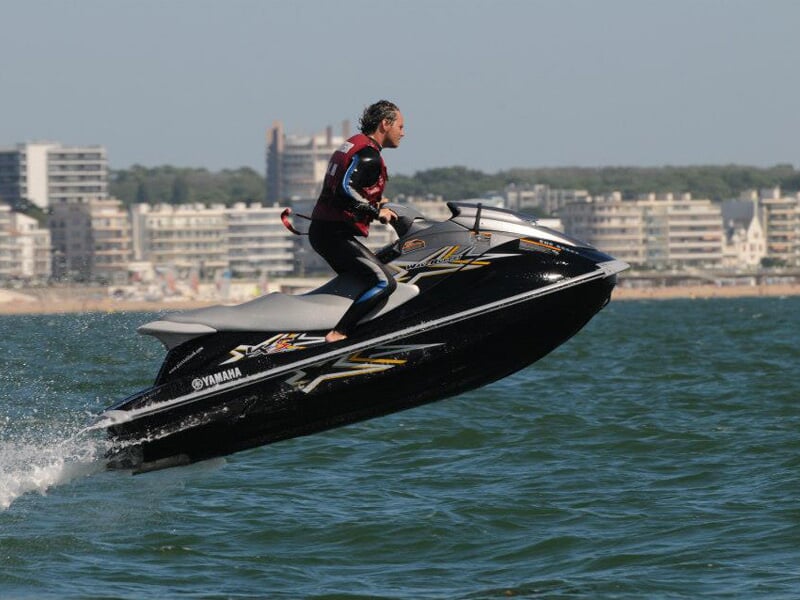 Homme en jet-ski Yamaha noir, effectuant un saut spectaculaire au-dessus de l’eau, avec des immeubles en arrière-plan