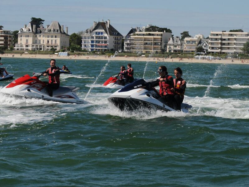 Groupe de personnes sur des jet-skis rouges et blancs, naviguant sur une mer agitée avec des bâtiments en arrière-plan