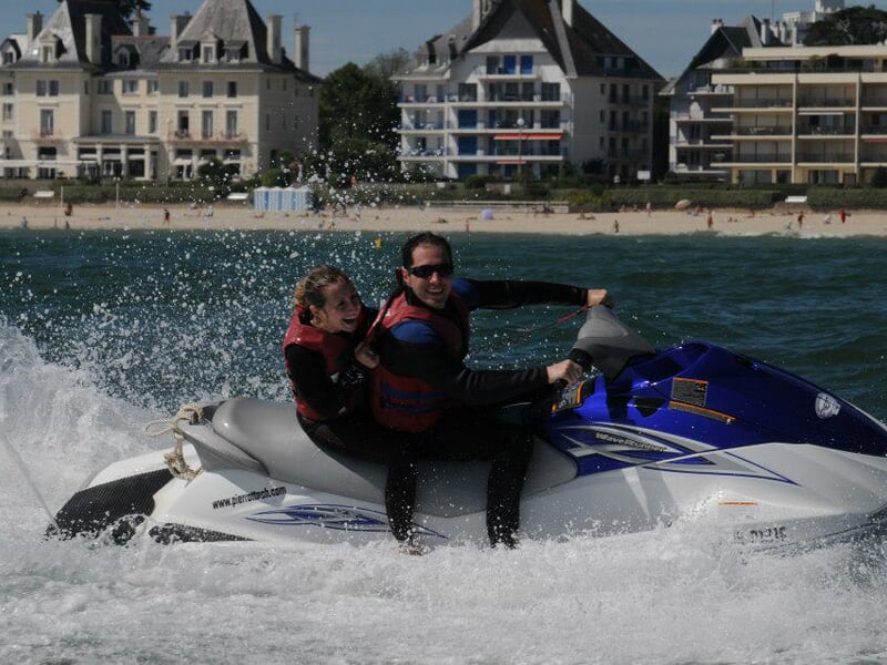 Couple en jet-ski Yamaha bleu, éclaboussures d’eau en action près de la plage bordée d’immeubles et de villas