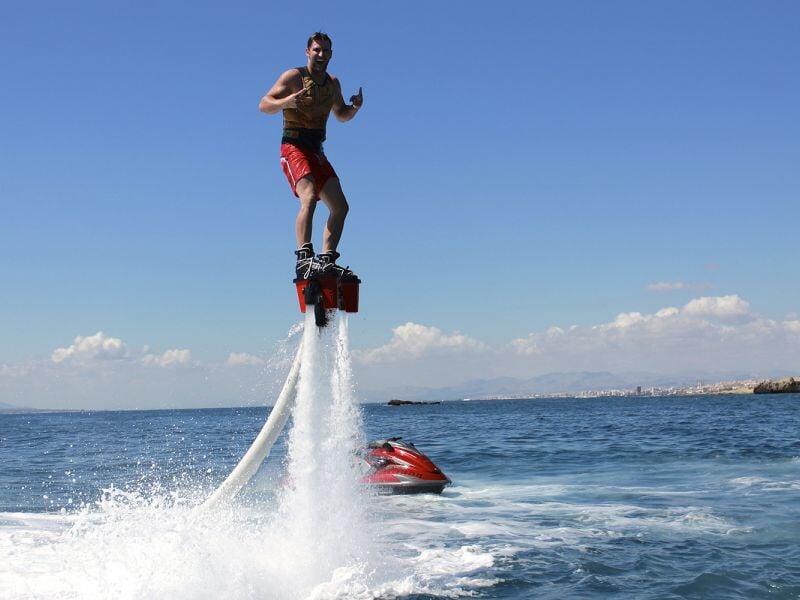 Homme en short rouge debout sur un flyboard propulsé par un jet d'eau au-dessus de la mer calme sous un ciel clair.