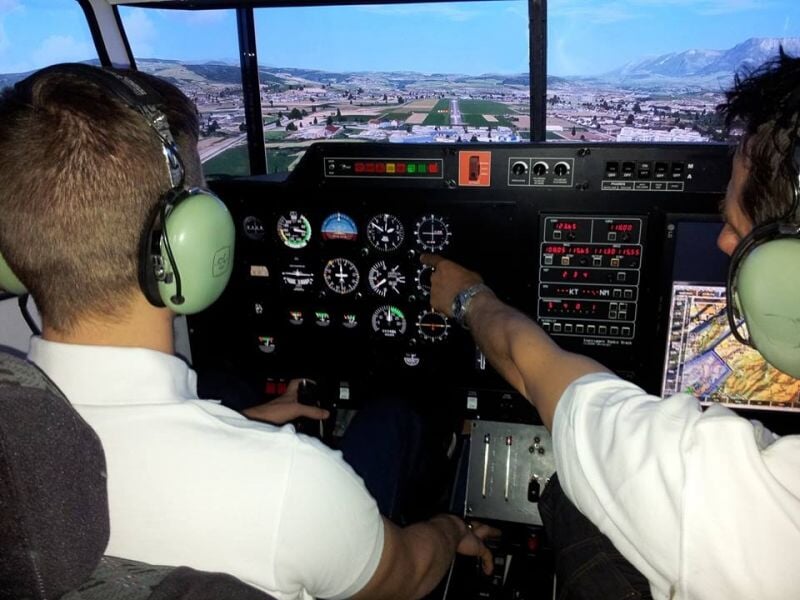 Deux personnes avec casque audio dans un simulateur de vol, l'une pointe vers les instruments du tableau de bord.