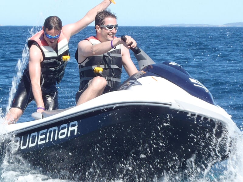 Deux personnes en gilets de sauvetage sur un jetski blanc et bleu naviguant sur une mer calme sous un ciel clair.