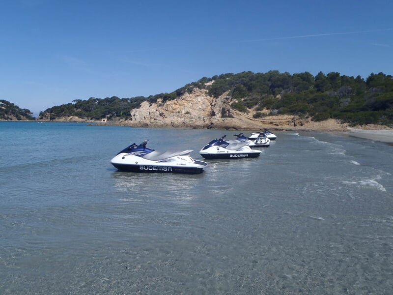 Quatre jetskis blancs et bleus alignés sur une plage de sable fin avec une mer calme et des collines en arrière-plan.