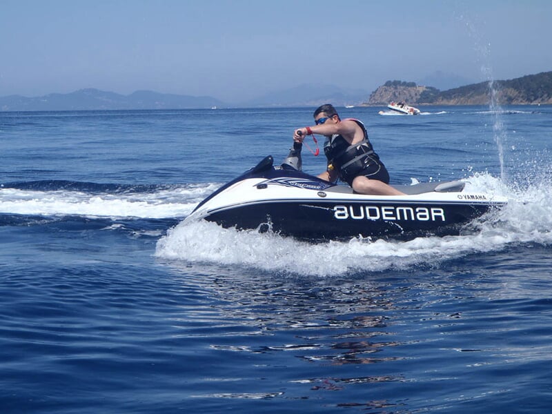Homme en gilet de sauvetage conduisant un jetski blanc et bleu sur une mer bleue avec des montagnes à l'horizon.