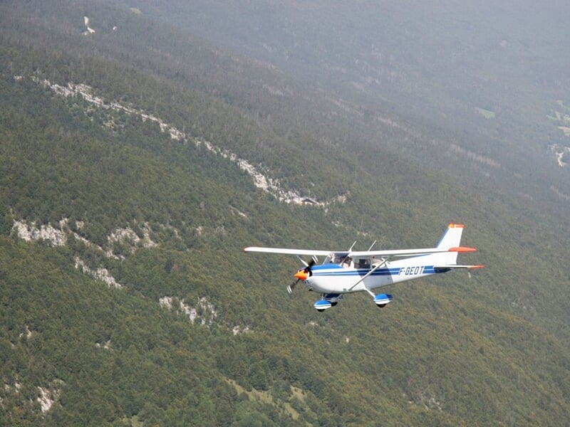 Avion léger blanc et bleu survolant une forêt dense avec des rochers visibles sur les pentes.