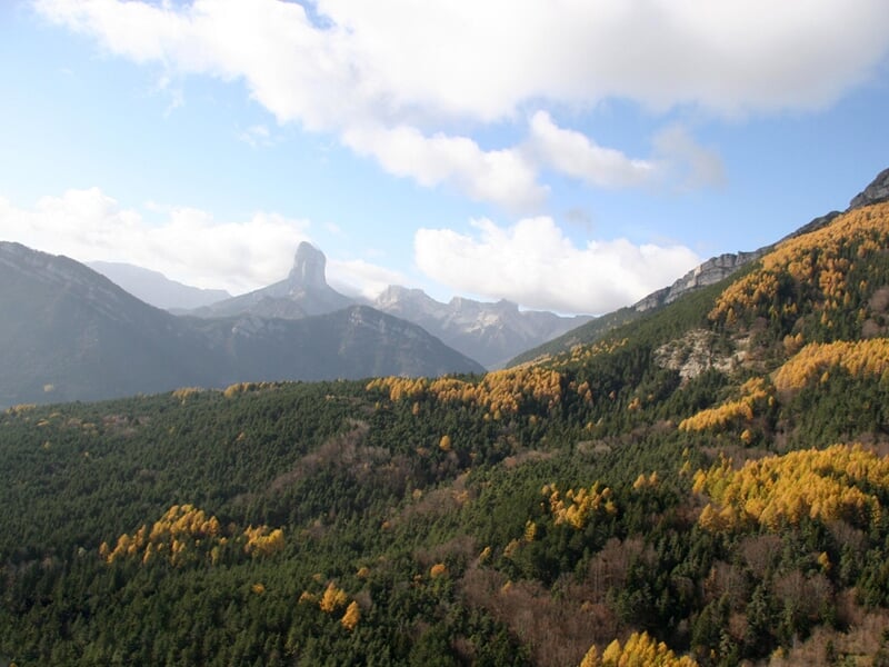 Paysage montagneux avec des arbres verts et jaunes sous un ciel partiellement nuageux en automne.