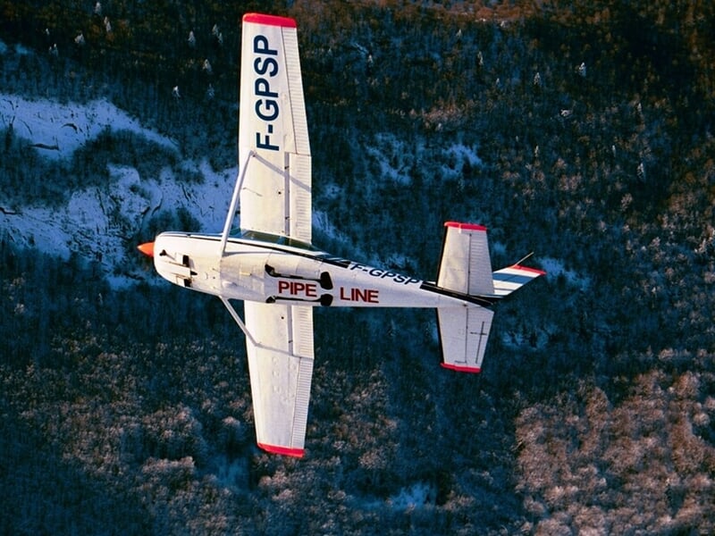 Avion blanc avec marquage rouge vu de dessus, volant au-dessus d'une forêt en partie enneigée.
