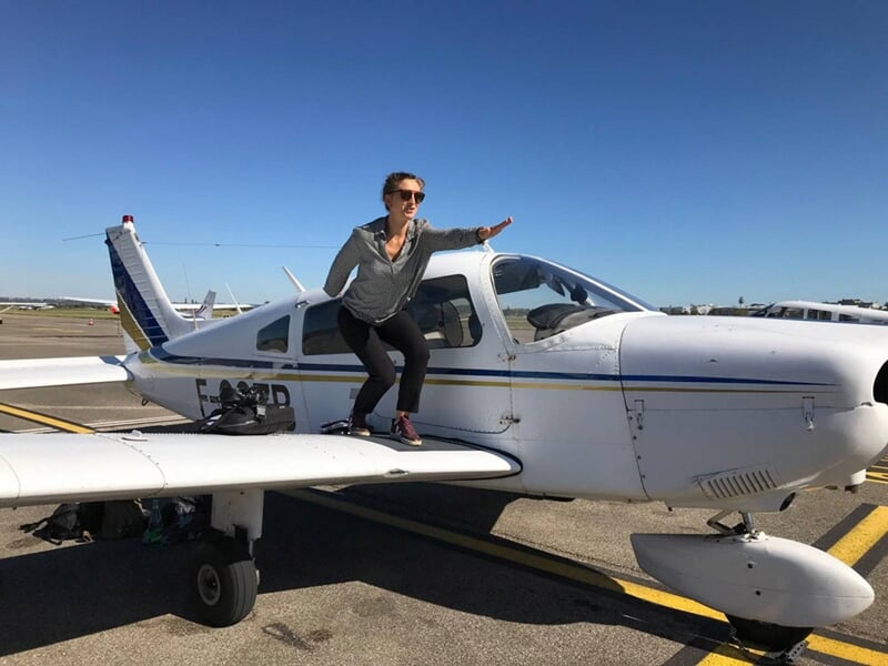 Personne posant debout sur l'aile d'un petit avion blanc stationné sur une piste d'aéroport par temps ensoleillé.