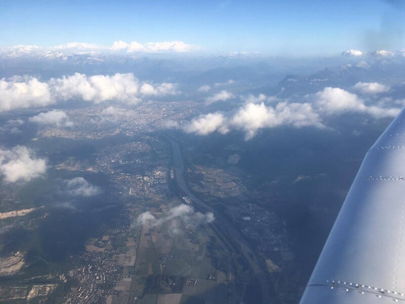 Vue aérienne d'un paysage avec des champs, une rivière et des collines partiellement couvertes de nuages blancs.