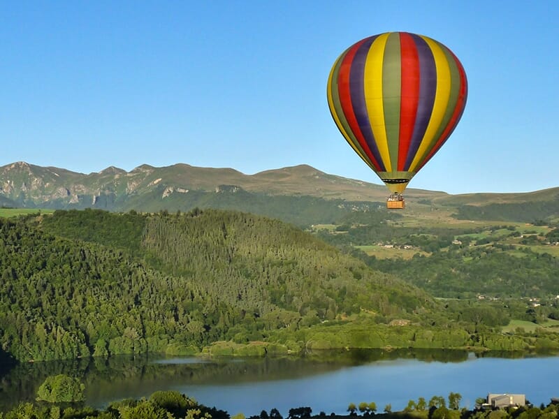 Montgolfière colorée volant au-dessus d'un paysage verdoyant avec un lac et des collines en arrière-plan.