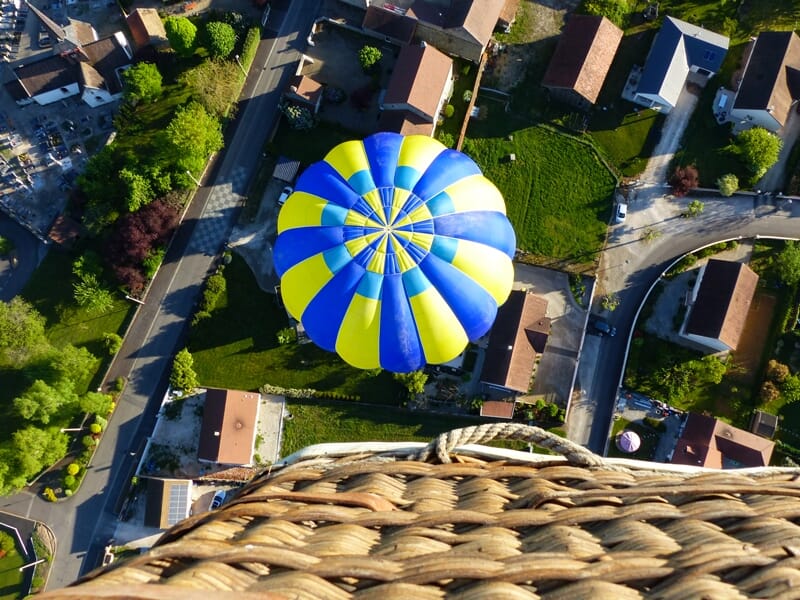 Vue aérienne d'une montgolfière bleue et jaune au-dessus d'un village avec des maisons et des routes.