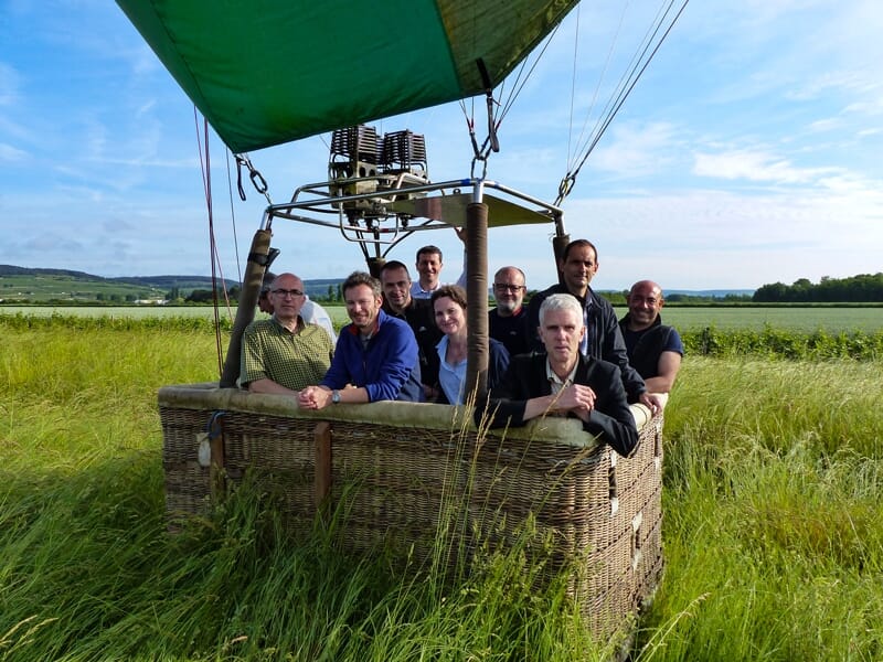 Groupe de personnes souriantes dans la nacelle d'une montgolfière prête à décoller dans un champ vert.
