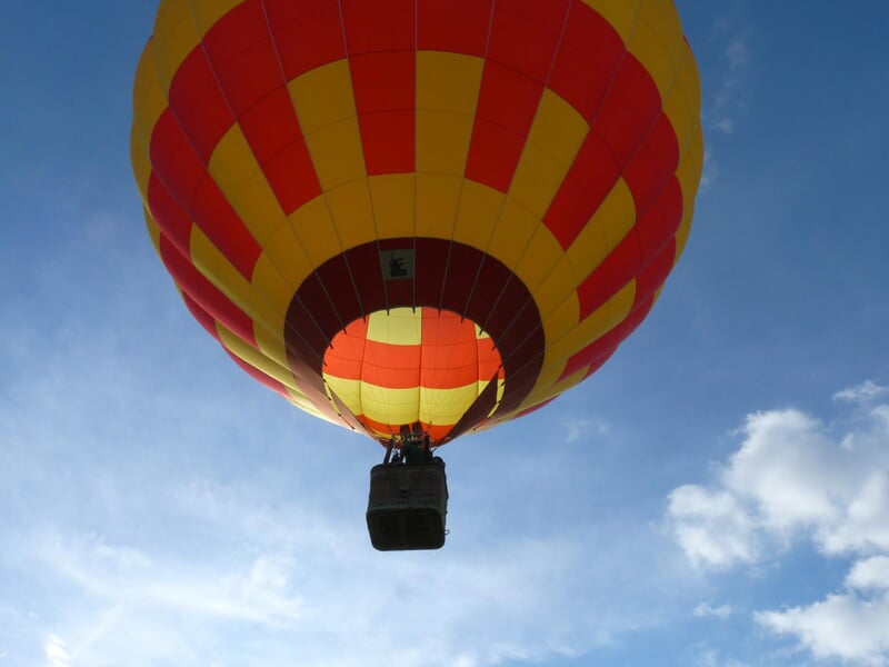Montgolfière rouge et jaune vue en contre-plongée, avec la nacelle suspendue sous un ciel bleu parsemé de nuages.