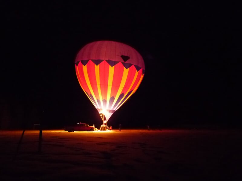 Montgolfière aux couleurs vives illuminée la nuit, gonflée par une flamme intense, avec une voiture et des personnes visibles à proximité sur un sol sombre.