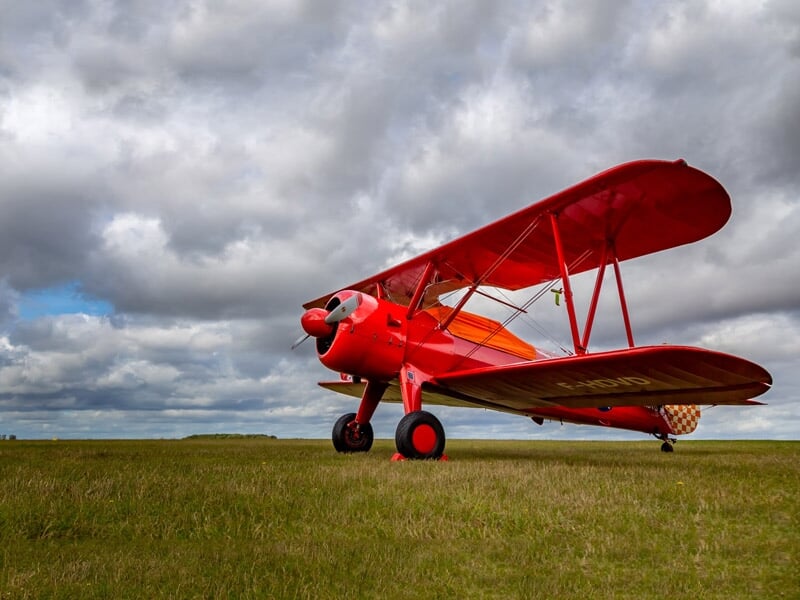 Avion rouge biplan posé sur un champ d'herbe verte sous un ciel nuageux, prêt pour un baptême de l'air.