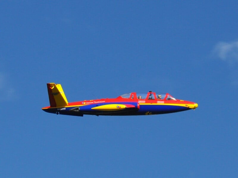 Un Fouga Magister rouge, bleu et jaune en vol sur un ciel bleu clair.