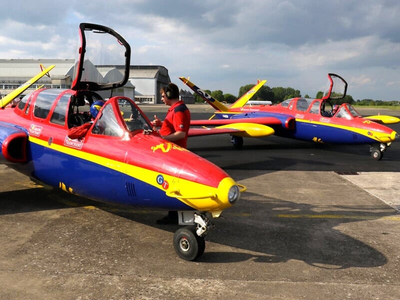 Des Fouga Magisters rouge, bleu et jaune alignés sur le tarmac avec des pilotes à côté.
