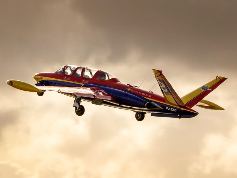 Un Fouga Magister rouge, bleu et jaune volant avec un ciel orageux en arrière-plan