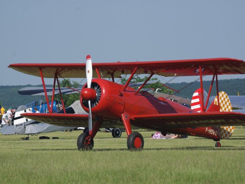 Avion rouge biplan stationné sur un terrain d'herbe avec plusieurs personnes et autres avions en arrière-plan.