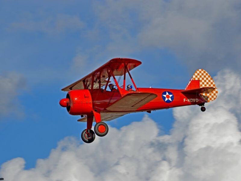 Avion rouge biplan en vol dans un ciel partiellement nuageux, effectuant une démonstration de voltige aérienne.