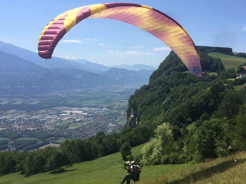 Vol en parapente biplace au-dessus d'une vallée verdoyante avec montagnes et village en arrière-plan sous un ciel bleu.