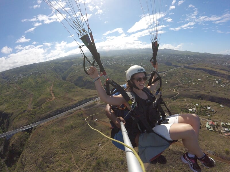 Femme souriante en vol en parapente biplace avec casque, tenant les commandes, paysage vallonné en dessous.