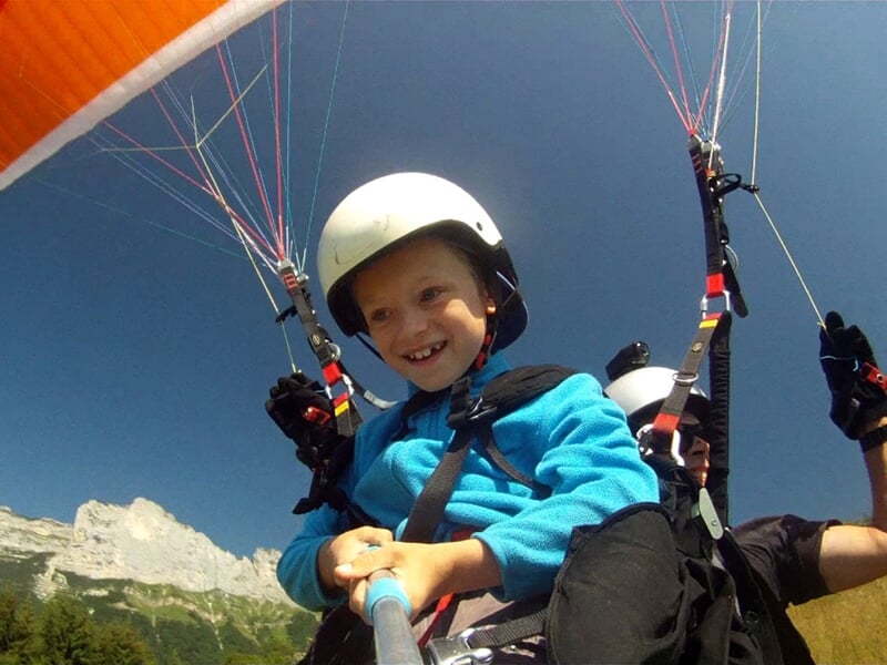 Enfant souriant en vol en parapente biplace avec casque, ciel bleu et montagnes en arrière-plan, prise selfie.