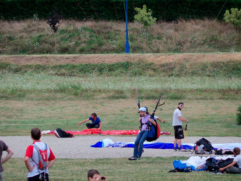 Personne en train de décoller en parapente sur un terrain herbeux avec plusieurs personnes autour en observation.