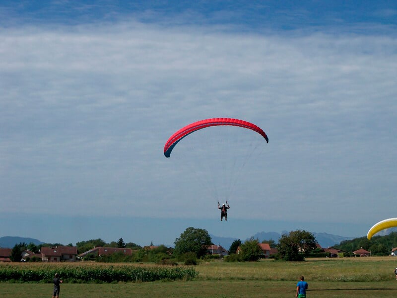 Parapentiste en vol au-dessus d'un champ vert avec un ciel bleu clair et quelques maisons en arrière-plan.