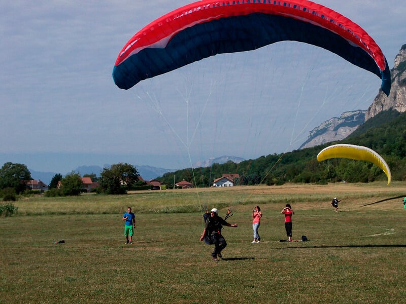 Atterrissage d'un parapentiste avec voile rouge et bleue sur un champ, plusieurs personnes regardent et montagnes en arrière-plan.